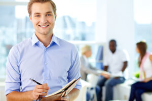 man in professional attire in office smiling and writing in notebook